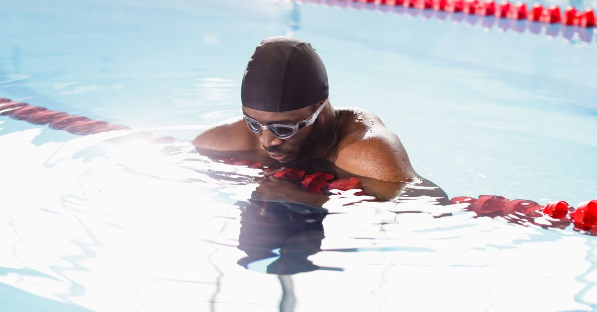 Man in a swimming pool with a black swimming cap on, holding onto the lane separators 